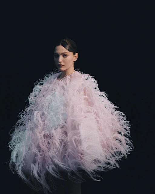 A striking portrait features a poised young woman in an extravagant, feathered pastel coat against a stark black background, studio lit.