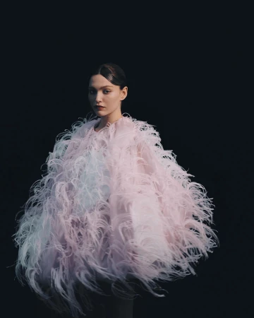 A striking portrait features a poised young woman in an extravagant, feathered pastel coat against a stark black background, studio lit.
