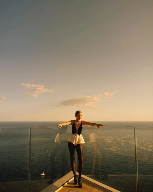 A female fashion model in a black and white outfit stands on a rooftop overlooking the ocean with clear blue sky in the background.