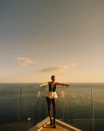 A female fashion model in a black and white outfit stands on a rooftop overlooking the ocean with clear blue sky in the background.