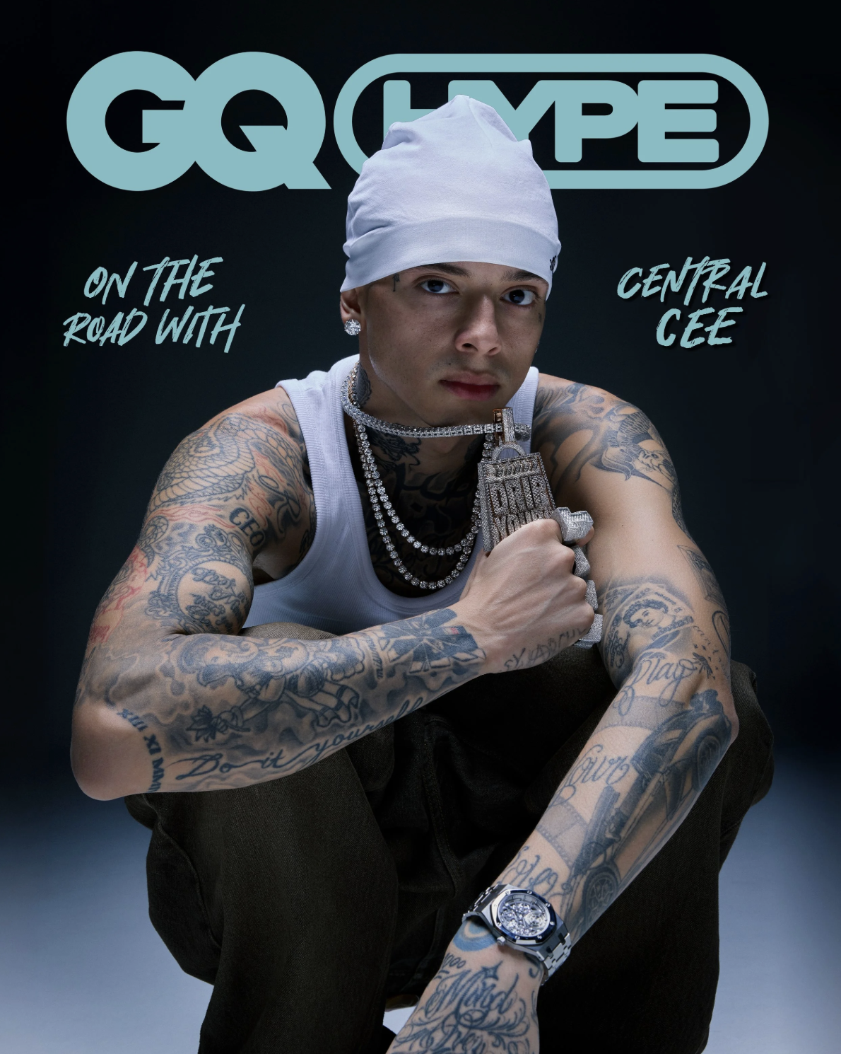 Central Cee wearing a white beanie and tank top with visible arm tattoos, holding a large diamond pendant against a dark background.