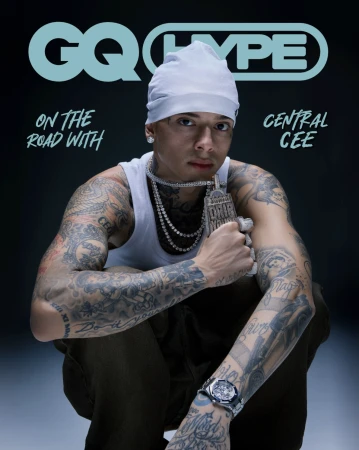 Central Cee wearing a white beanie and tank top with visible arm tattoos, holding a large diamond pendant against a dark background.