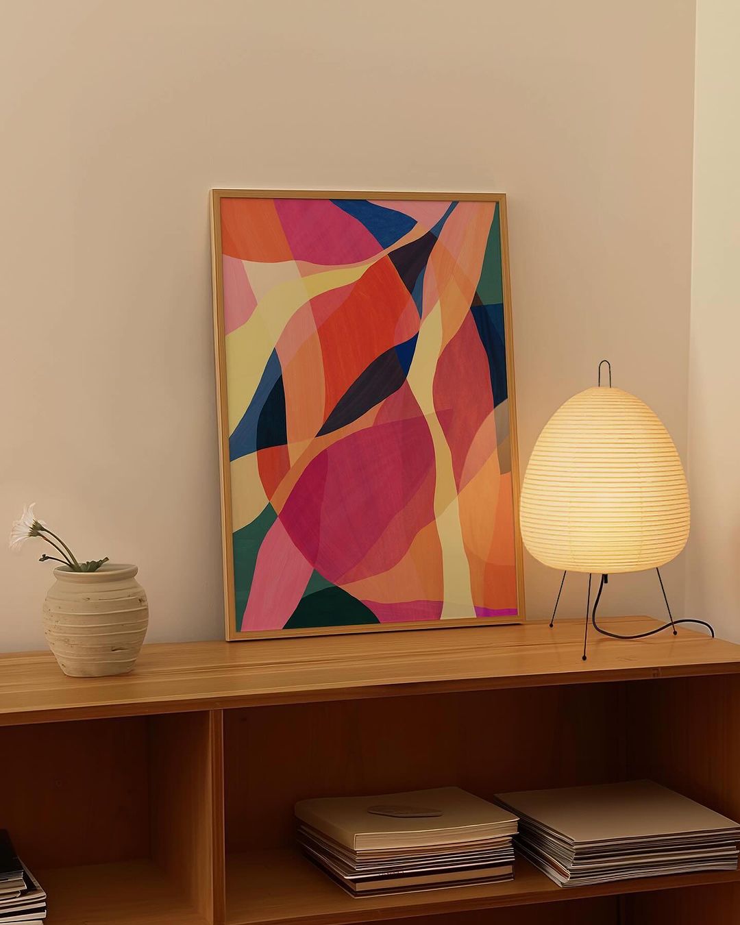 Abstract art print with wood frame displayed above shelf with books, next to a paper globe lamp in a neutral-toned room.