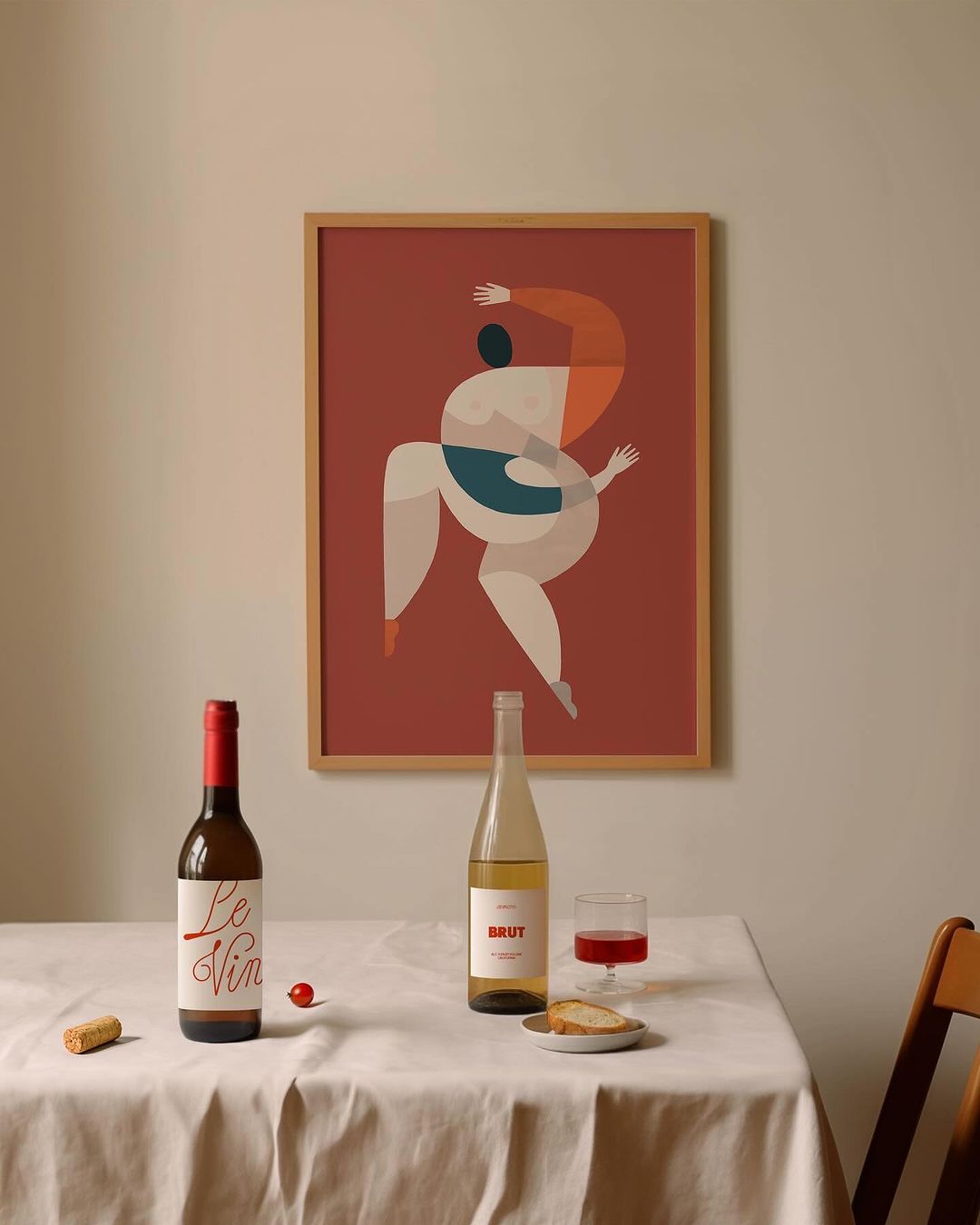 Still life featuring wine bottles, a glass, and a framed art print of a figure on a tablecloth-covered table in an indoor setting.