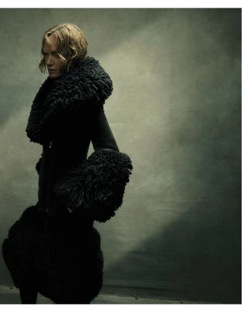 Woman wearing a heavy black textured coat with voluminous faux fur collar and cuffs against a muted studio background.