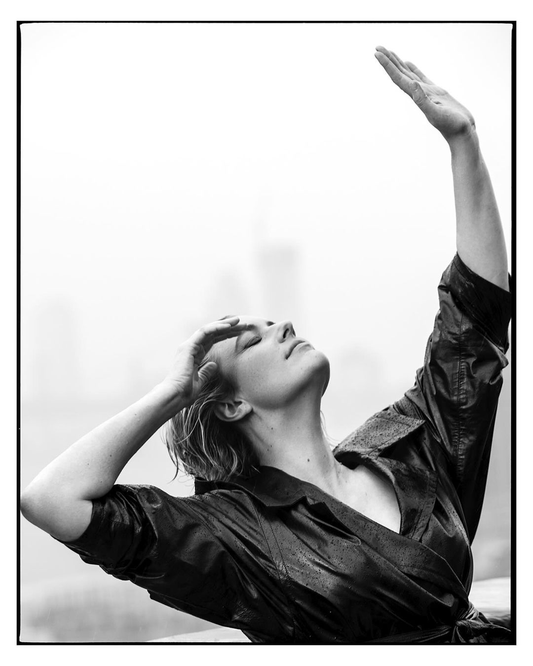 Monochrome portrait of a woman in a shiny coat with her hands raised, looking up with a blurred city background.
