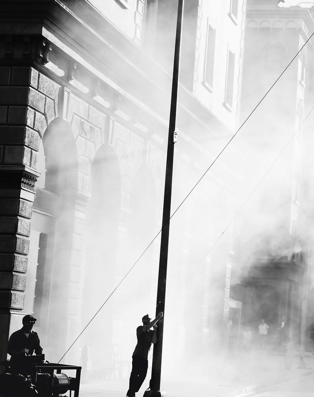 Black and white photograph of a man leaning against a pole in a misty outdoor setting, with a second man nearby and a generator.