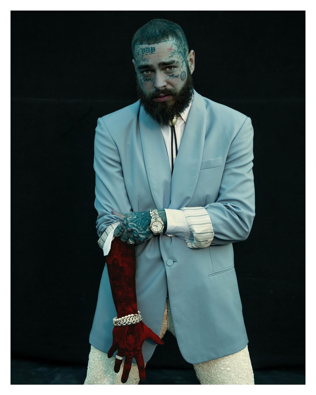 Post Malone poses in a light blue suit with tattoos visible on his face and arms against a dark background, wearing jewelry and a bolo tie.