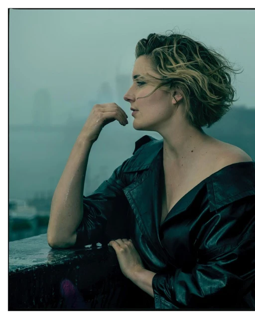 A woman with short blonde hair in a black leather jacket poses in profile on a rooftop with a misty cityscape in the background.