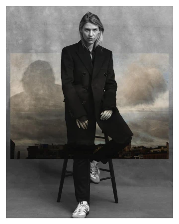 Studio shot of a woman with fair skin in a black double-breasted coat and black pants, sitting on a wooden stool in front of a painted backdrop.