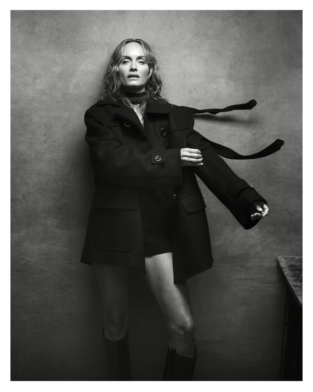 Monochrome portrait of a woman with long hair in an open black coat, mini shorts, and boots, standing against a textured grey background.