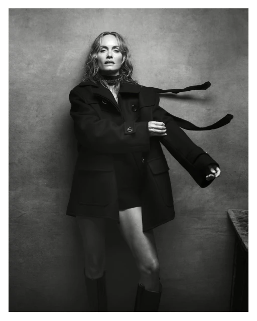Monochrome portrait of a woman with long hair in an open black coat, mini shorts, and boots, standing against a textured grey background.
