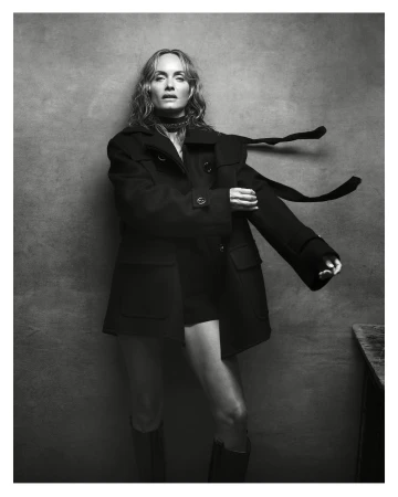 Monochrome portrait of a woman with long hair in an open black coat, mini shorts, and boots, standing against a textured grey background.