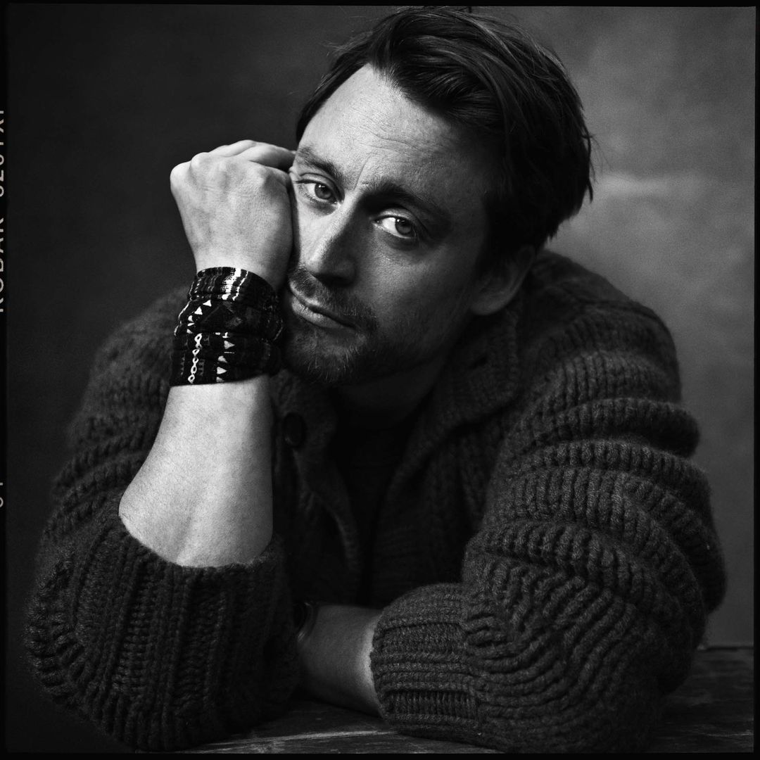 Black-and-white close-up portrait of a pensive adult male with a hand on his face, wearing a sweater and a studded bracelet.