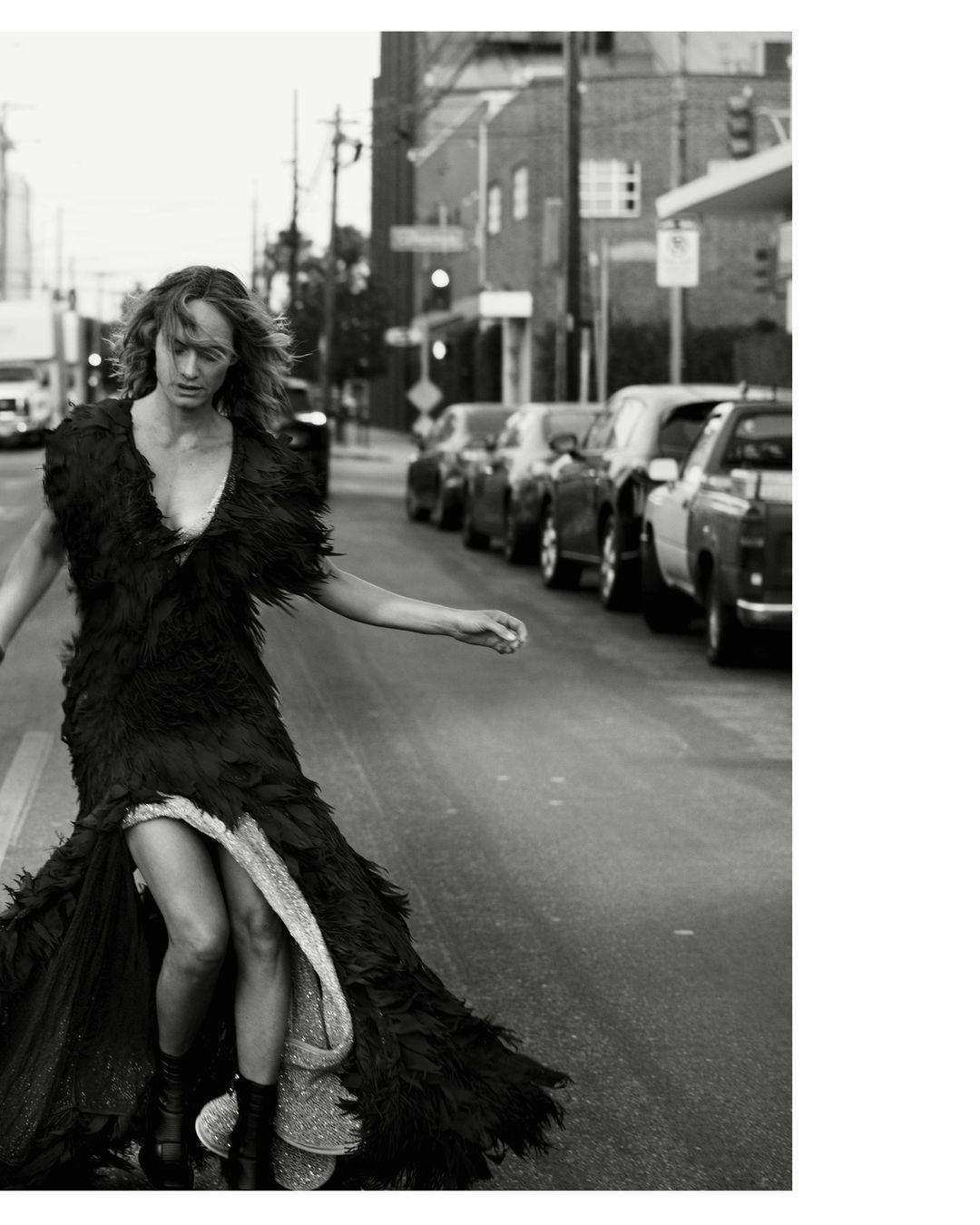 Monochrome fashion shot of a woman in a feathered gown walking on an urban street lined with cars and buildings in a grayscale photo.