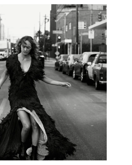 Monochrome fashion shot of a woman in a feathered gown walking on an urban street lined with cars and buildings in a grayscale photo.