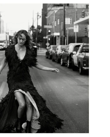 Monochrome fashion shot of a woman in a feathered gown walking on an urban street lined with cars and buildings in a grayscale photo.