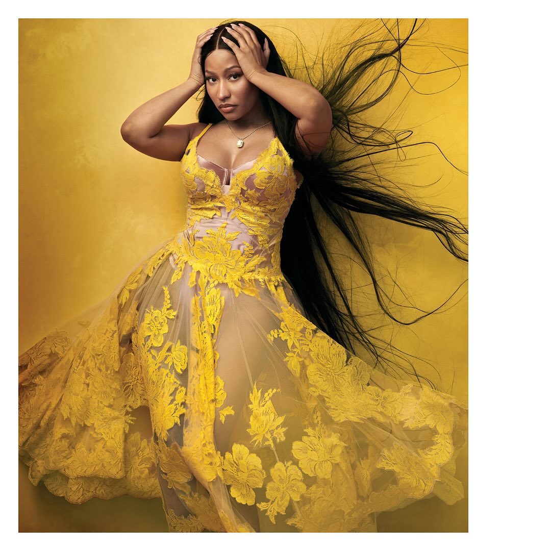 Portrait of Nicki Minaj in a flowing yellow lace dress, standing against a yellow background with long black hair styled in the wind.