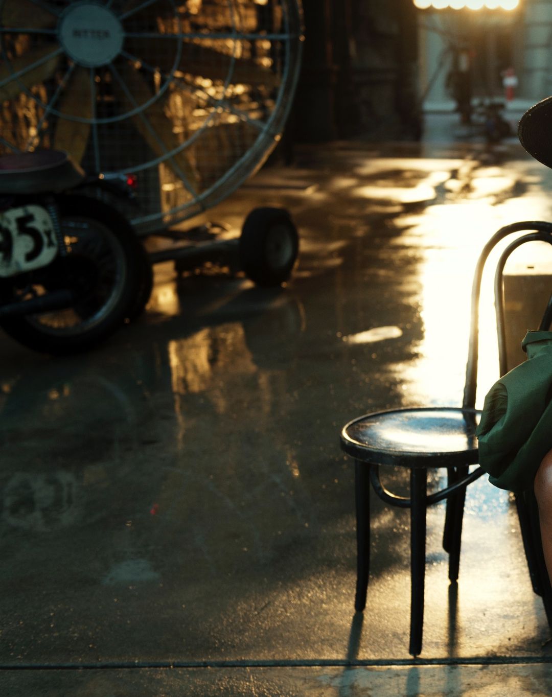 Indoor scene with a large industrial fan, small motorcycle, and a person partially visible sitting on a chair, warm color grading.