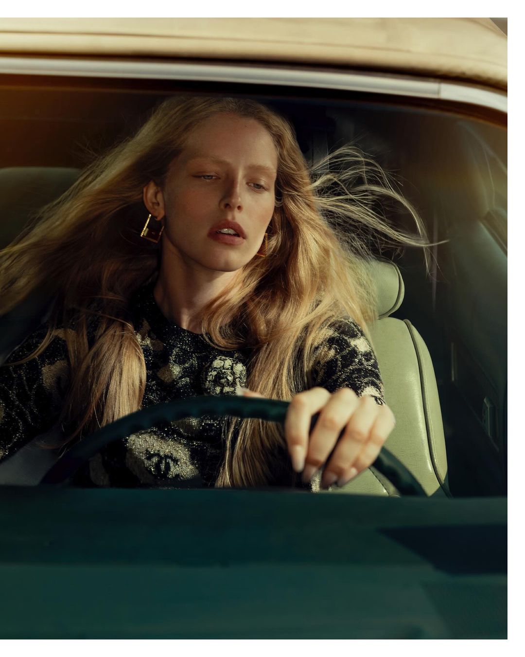 Portrait of a blonde woman driving a car, wearing a patterned black top and gold earrings, medium shot with warm light.