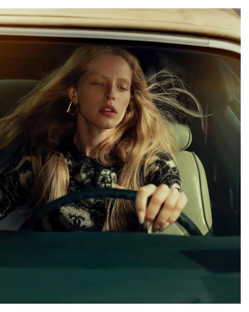 Portrait of a blonde woman driving a car, wearing a patterned black top and gold earrings, medium shot with warm light.
