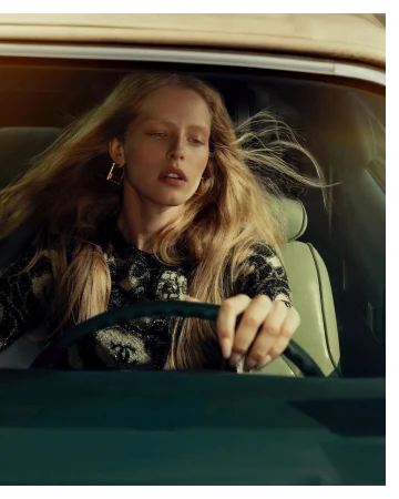 Portrait of a blonde woman driving a car, wearing a patterned black top and gold earrings, medium shot with warm light.