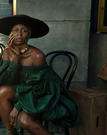 Elegant Black woman in a green dress and large hat poses on a chair in an indoor setting. Gold jewelry and nail art add to her refined style.