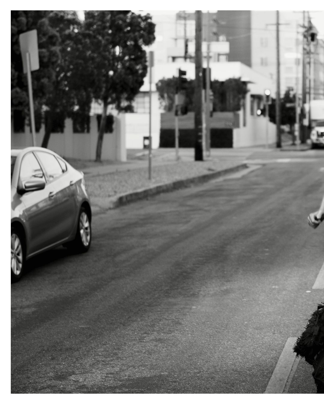 Black and white shot of a car on a road next to a curb, with a glimpse of a person on the right side of the frame.