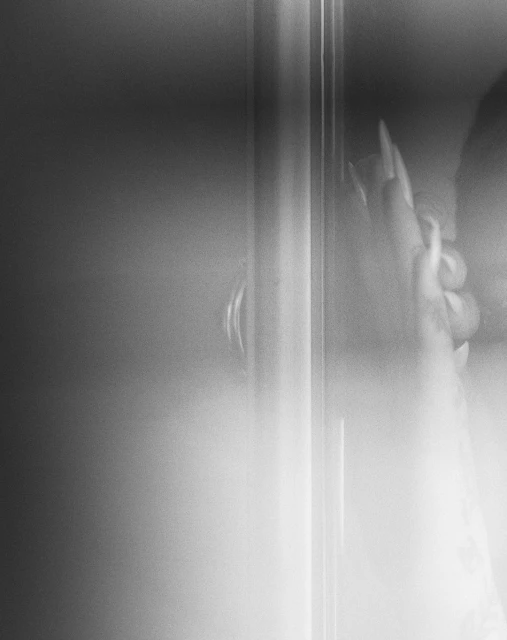 Close-up of a person's hand with long nails gripping a metal bar; the image is black and white with a soft, blurred effect.