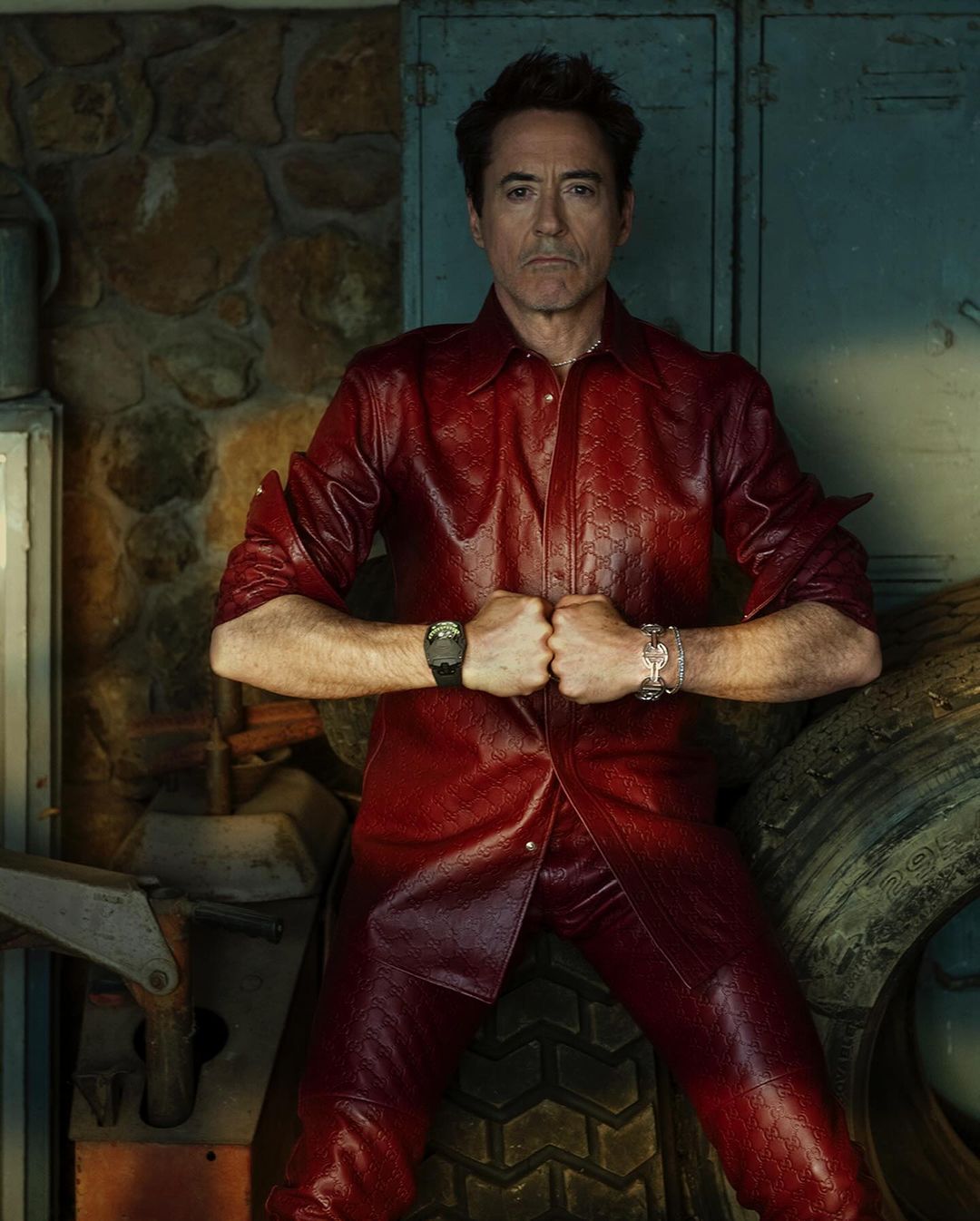 Close-up of adult man in red textured outfit with garage door and stone wall background, arms flexed, wearing a watch and bracelet.