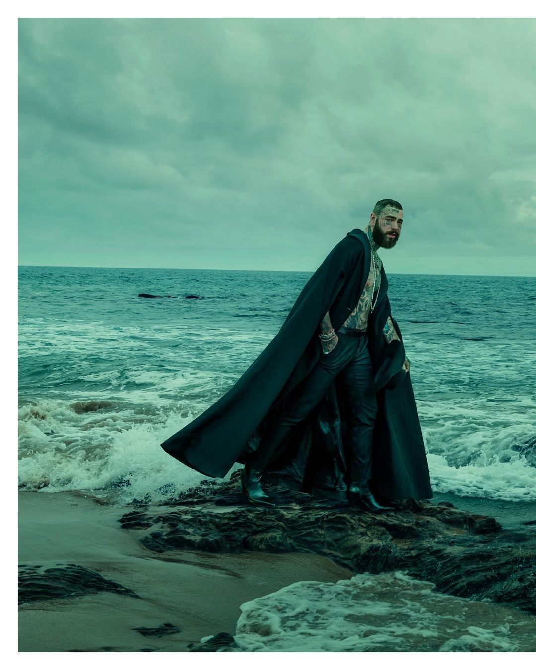 A bearded man in a long black cape stands on a rocky shore with the ocean and sky in the background in a captivating outdoor portrait.