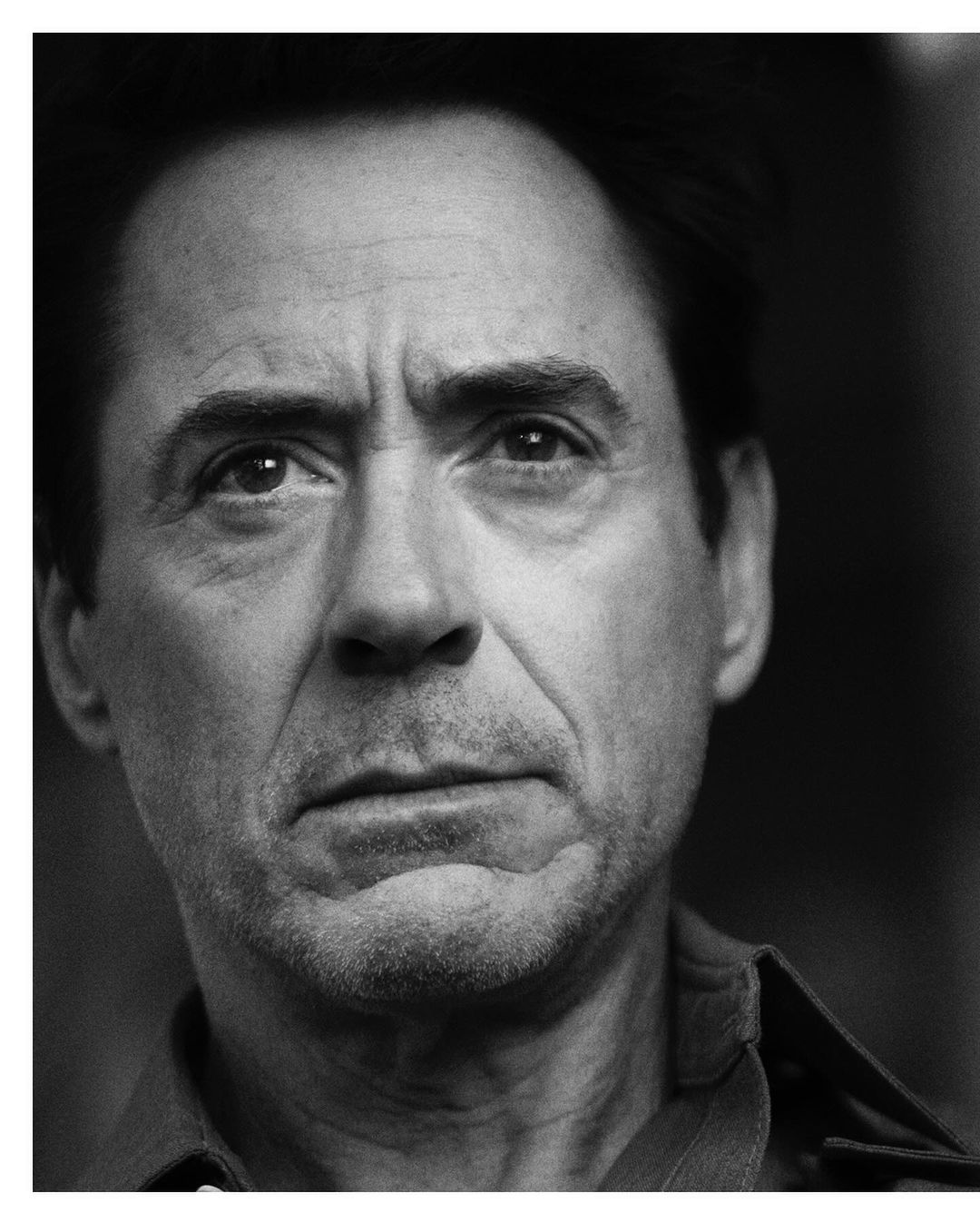 Close-up black and white portrait of an adult male with a serious expression, wearing a collared shirt, captured in a photo studio setting.