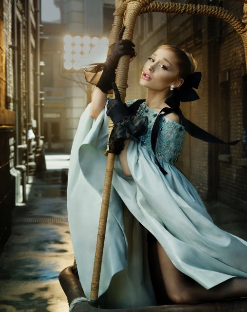 Adult female with black gloves poses holding onto rope in an urban setting, wearing an ice-blue dress and pearl earrings with hair up in a bow.