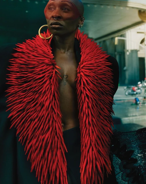 Dramatic portrait of a bald adult wearing a vibrant red scarf-like garment and gold hoops outdoors in an urban setting. Fashion editorial.