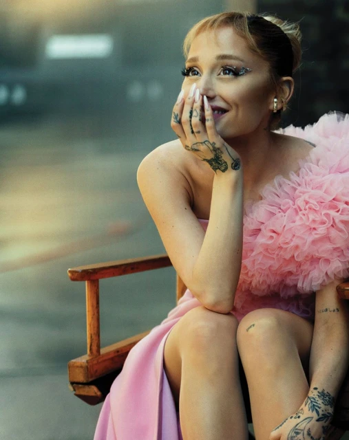 Smiling woman in a pink dress with elaborate eye makeup sits on a director's chair with tattoos on her hands and arms.