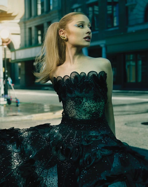 Elegant woman in black strapless gown poses on urban street, showcasing a high ponytail, gold jewelry, and sophisticated makeup for a high-fashion vibe.