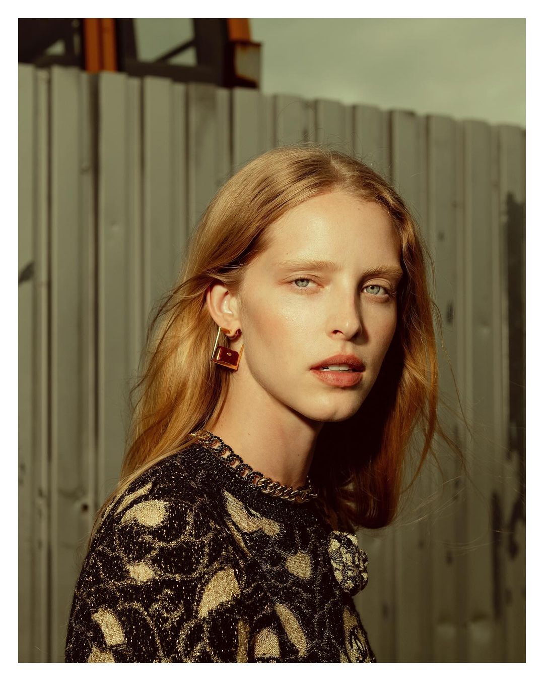 Close-up portrait of a fair-skinned woman with light blonde hair, wearing statement earrings and a patterned top against a neutral backdrop.