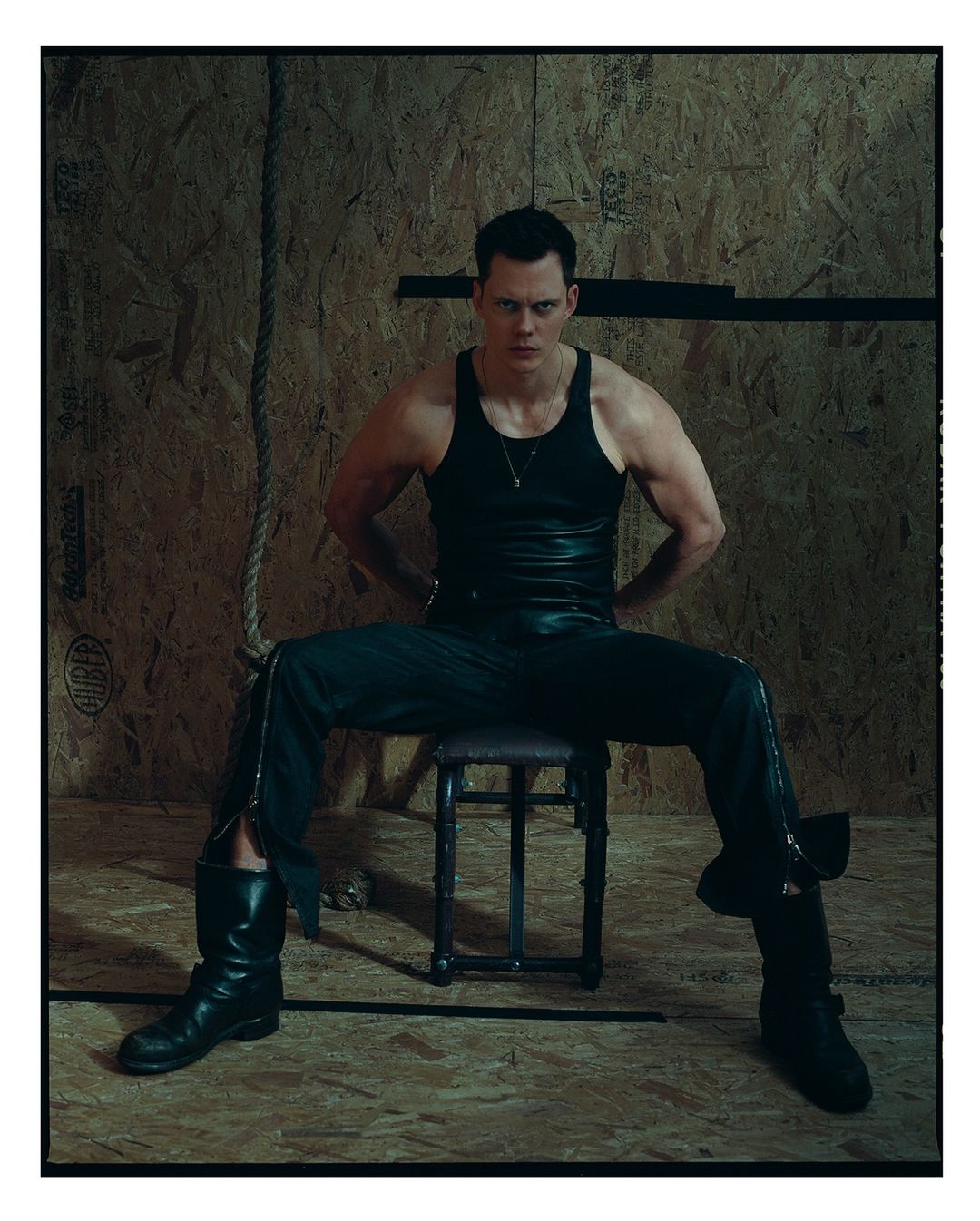 Portrait of an adult male wearing a black tank top, dark pants, and boots, sitting on a stool against a plywood wall background.