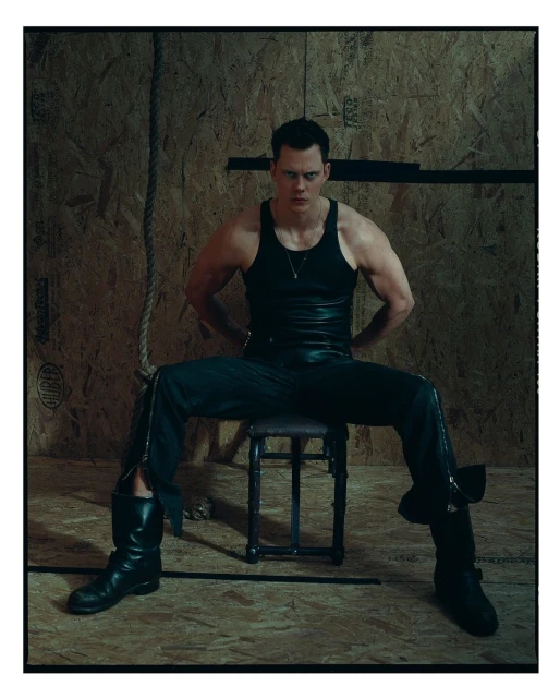 Portrait of an adult male wearing a black tank top, dark pants, and boots, sitting on a stool against a plywood wall background.
