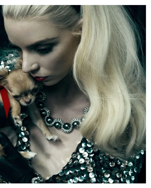 Adult female holding chihuahua, looking down with closed mouth, indoors. The woman wears a sequined top and necklace.