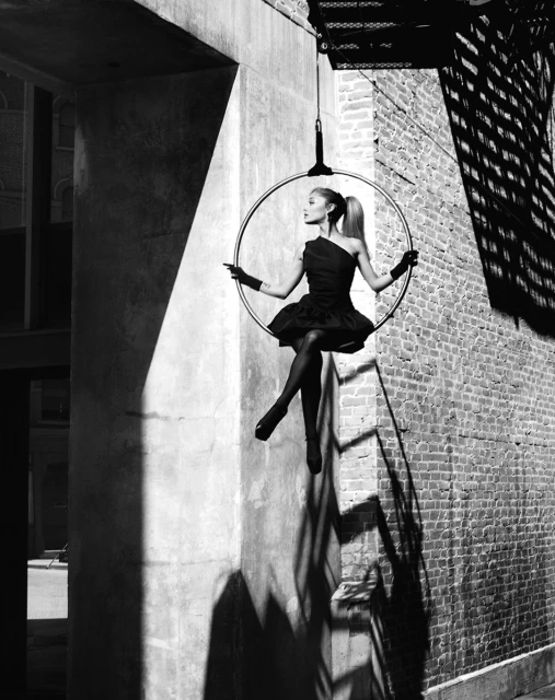 Black-and-white shot of a woman in a dark dress and tights performing aerial acrobatics on a hoop suspended between buildings.