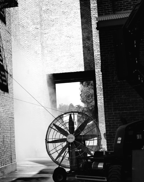 Industrial fan with large blades positioned between brick buildings, creating a cooling or atmospheric effect in monochrome.