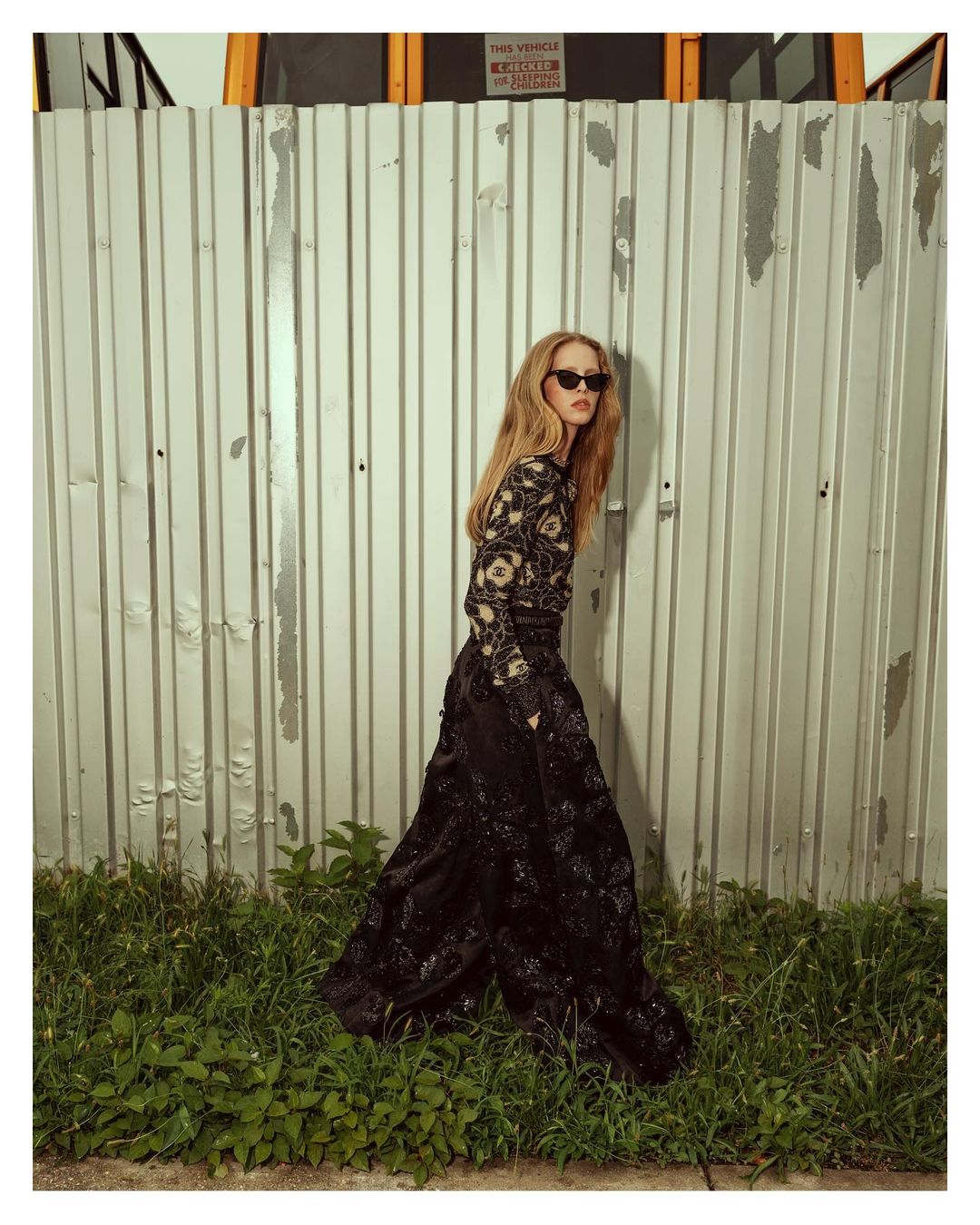 A slender woman in a black dress with floral accents stands against a corrugated metal wall, complemented by green grass at the bottom.
