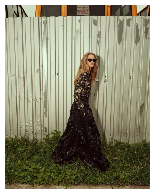 A slender woman in a black dress with floral accents stands against a corrugated metal wall, complemented by green grass at the bottom.