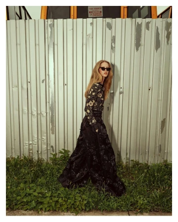 A slender woman in a black dress with floral accents stands against a corrugated metal wall, complemented by green grass at the bottom.
