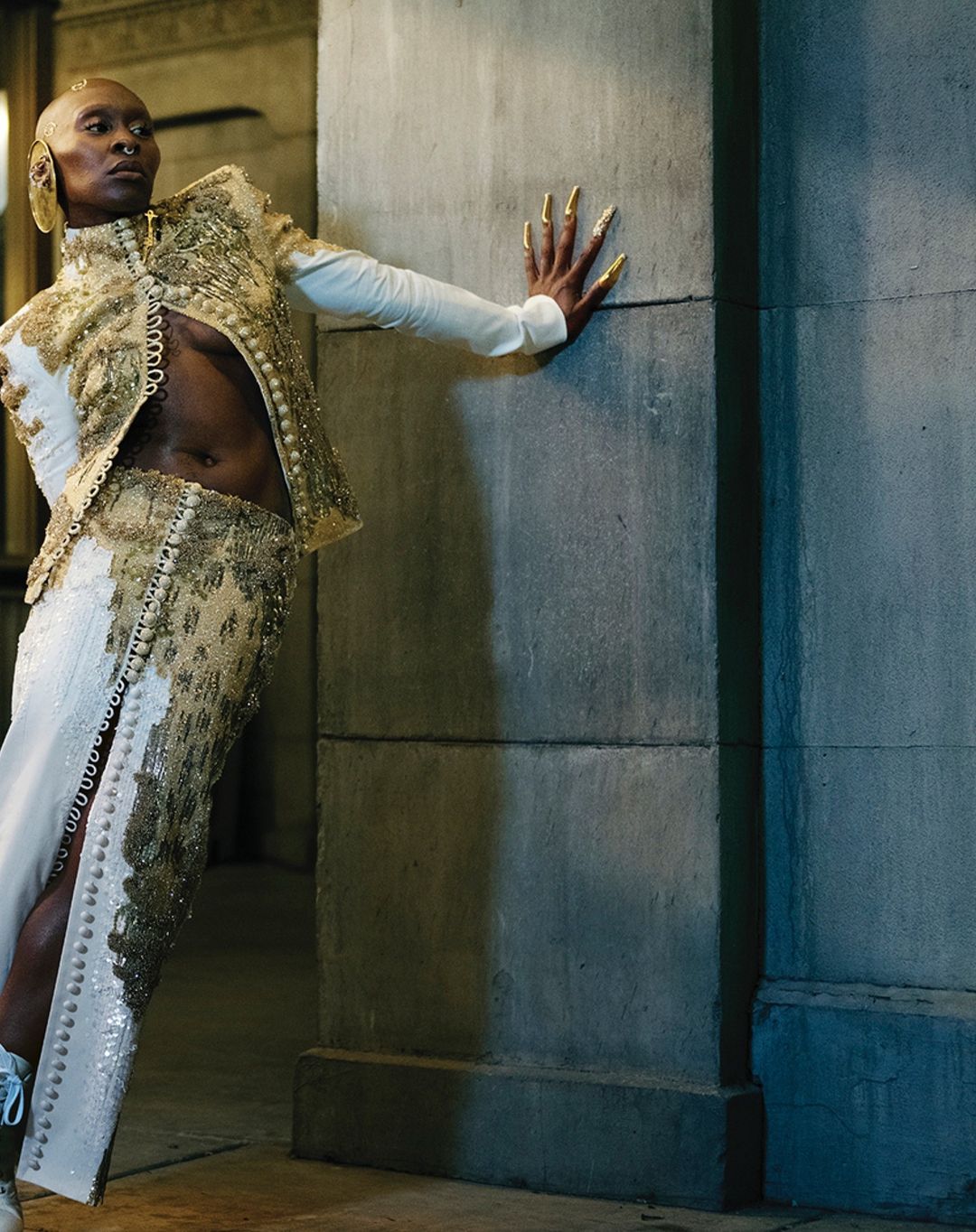 Elegant adult in a custom white and gold beaded suit, stands against a grey concrete wall with sunlight. Unique fashion portrait.