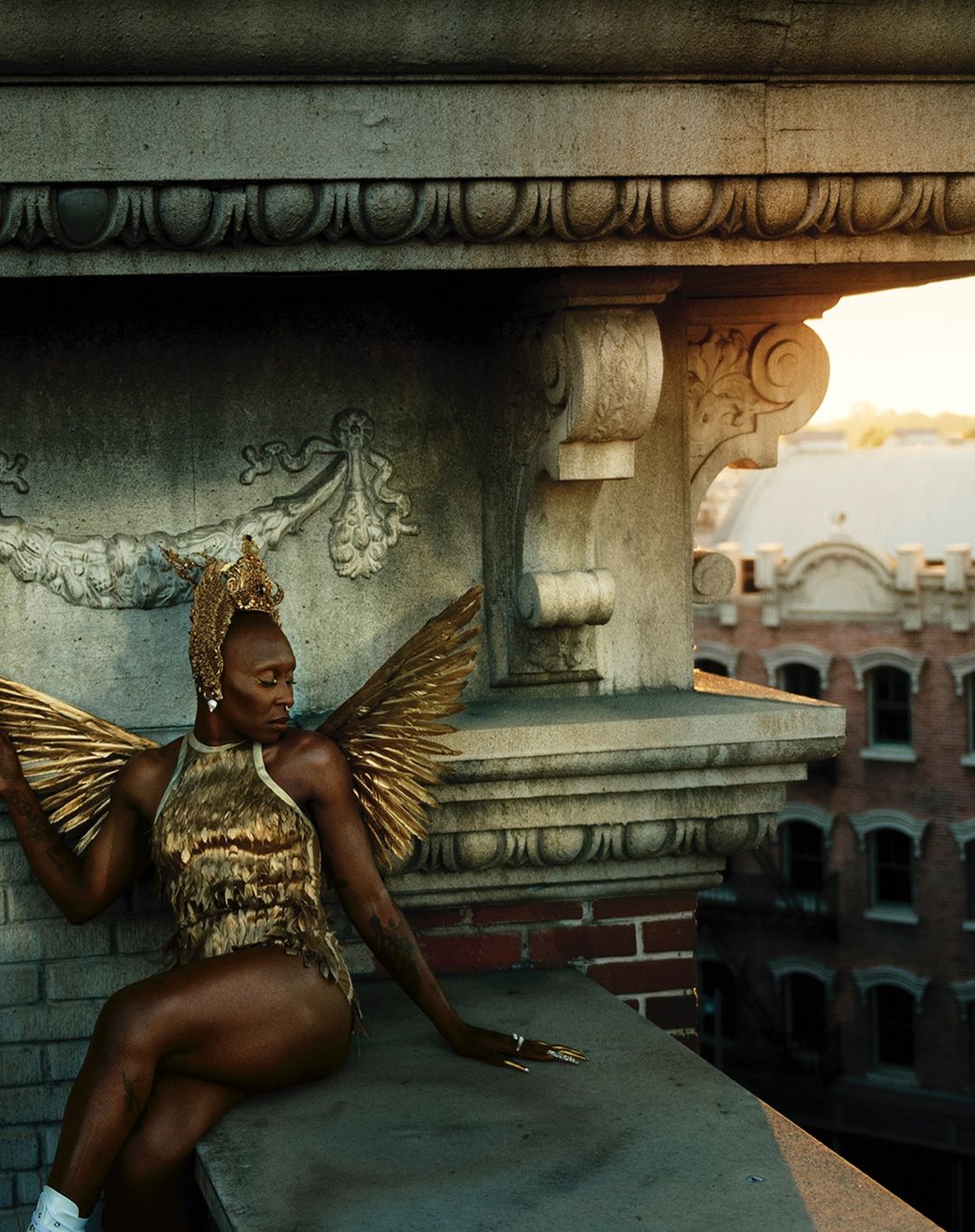 Woman in gold winged costume sitting on building ledge with city view. Dramatic rooftop fashion portrait with architectural details and urban scene.