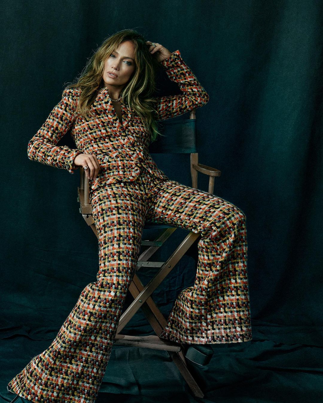 Jennifer Lopez sits on a director's chair, in a patterned tweed suit and bell-bottom pants in a beauty and fashion portrait.
