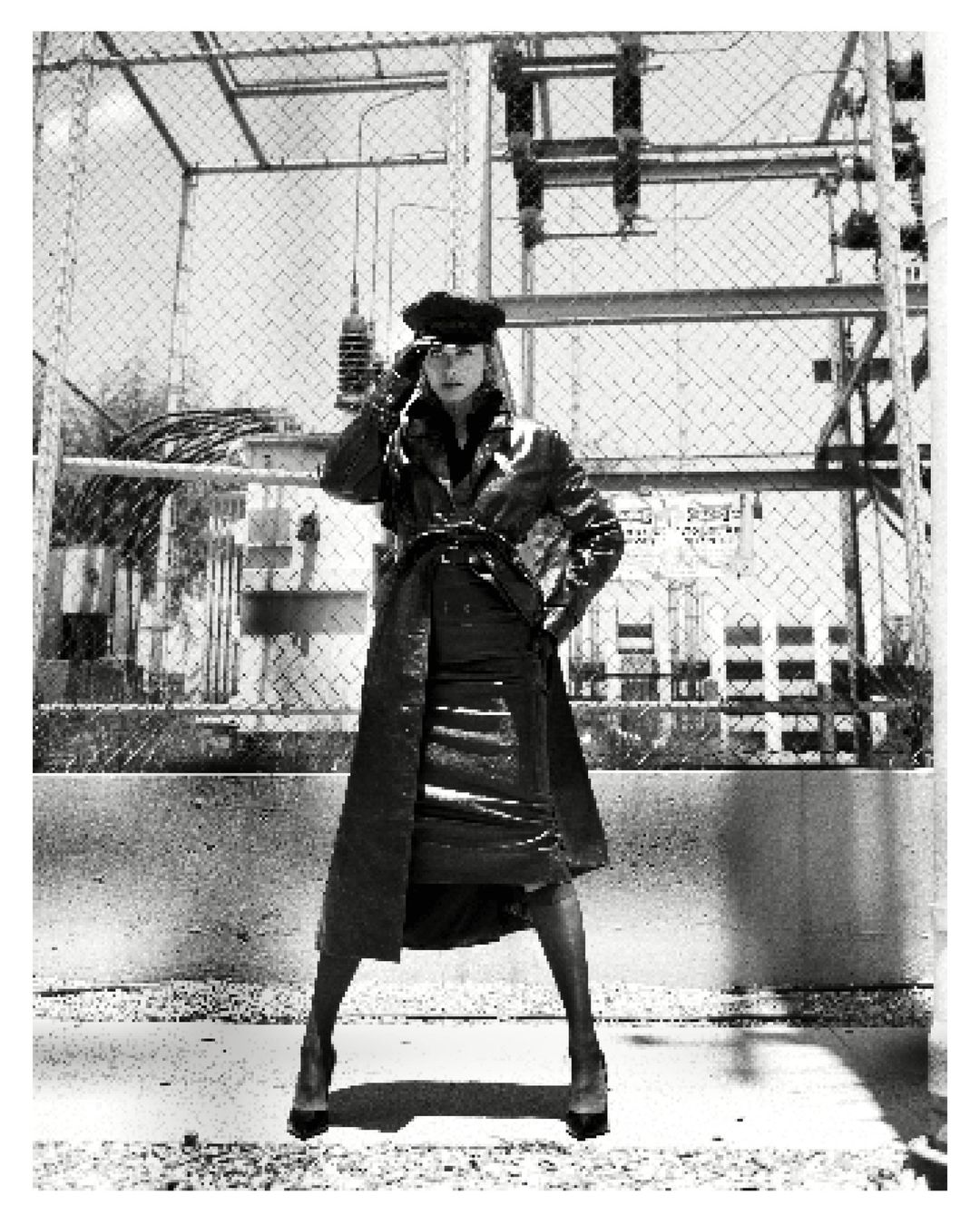 Monochrome fashion shot of a woman wearing a long coat and hat posing outdoors against an industrial background with a fence.