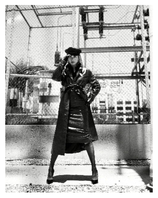 Monochrome fashion shot of a woman wearing a long coat and hat posing outdoors against an industrial background with a fence.
